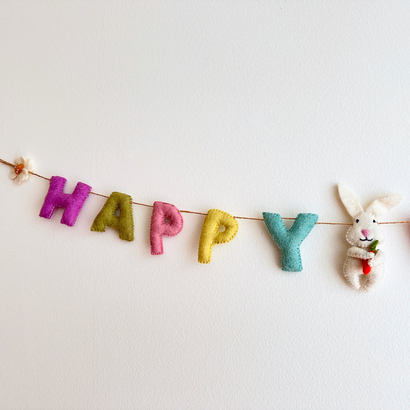 A pastel-toned Happy Easter felt garland is displayed against a plain white background. The wool felt garland spells "HAPPY" with individual capital letters in pastel purple, light green, pink, and yellow. A white felt bunny holding a small orange carrot hangs next to "HAPPY". The garland is strung on durable jute thread, ending with a white daisy.