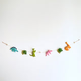 The vertical Jurassic dinosaur felt garland is displayed against a bright white wall, featuring a playful arrangement of felt shapes. It includes a blue stegosaurus, green monstera leaf, green T-Rex, pink triceratops, another green monstera leaf, and an orange brontosaurus. A small copper bell dangles from the bottom, adding a whimsical touch.