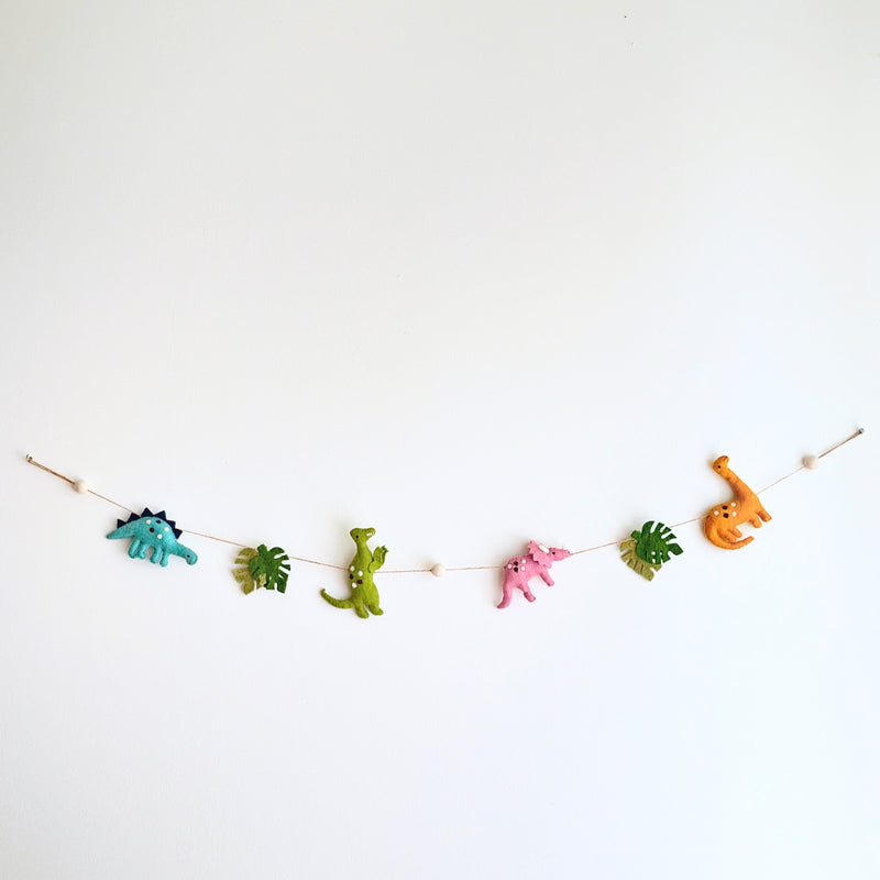 The vertical Jurassic dinosaur felt garland is displayed against a bright white wall, featuring a playful arrangement of felt shapes. It includes a blue stegosaurus, green monstera leaf, green T-Rex, pink triceratops, another green monstera leaf, and an orange brontosaurus. A small copper bell dangles from the bottom, adding a whimsical touch.
