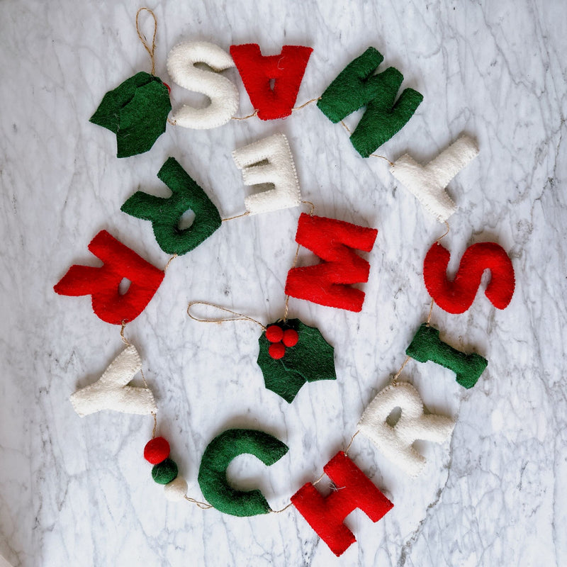 A 'Merry Christmas' wool felt garland is displayed in a semi-circular arrangement on a marbled white surface. This handcrafted garland features individual, soft wool felt letters spelling 'Merry Christmas' in alternating red, green, and white. It is strung on natural twine and accented with decorative green felt holly leaves and red felt berries, creating a festive and cheerful display.