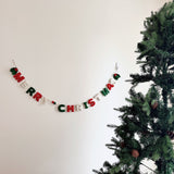 A festive 'Merry Christmas' felt garland hangs horizontally on a white wall, positioned next to a decorated Christmas tree. The hand-stitched letters, crafted from classic red, green, and white felt, spell 'Merry Christmas.' Small green felt holly leaves and red berries adorn the garland's ends, adding traditional charm to the holiday display.
