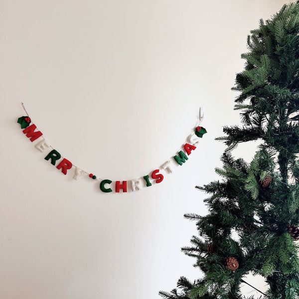 A festive 'Merry Christmas' felt garland hangs horizontally on a white wall, positioned next to a decorated Christmas tree. The hand-stitched letters, crafted from classic red, green, and white felt, spell 'Merry Christmas.' Small green felt holly leaves and red berries adorn the garland's ends, adding traditional charm to the holiday display.