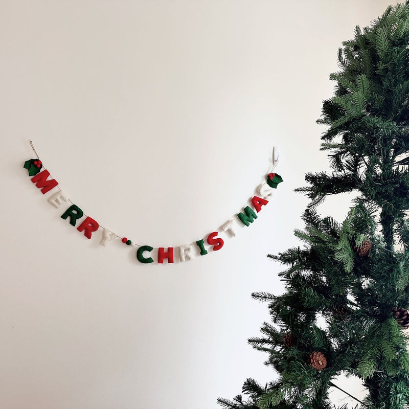 A festive 'Merry Christmas' felt garland hangs horizontally on a white wall, positioned next to a decorated Christmas tree. The hand-stitched letters, crafted from classic red, green, and white felt, spell 'Merry Christmas.' Small green felt holly leaves and red berries adorn the garland's ends, adding traditional charm to the holiday display.