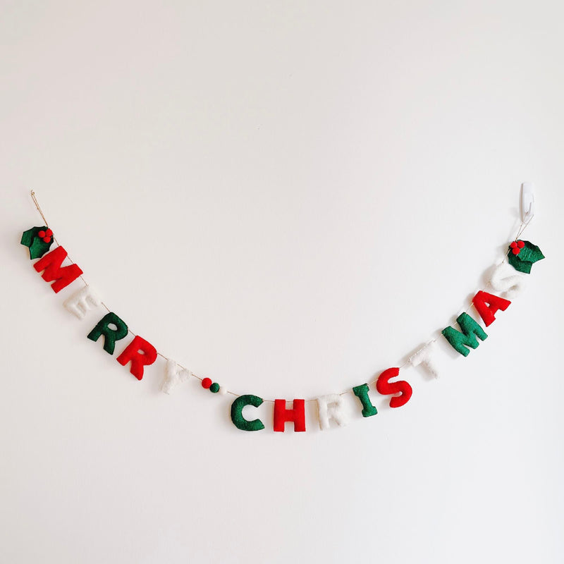 A felt Merry Christmas garland is hung against a plain white wall, suspended in a gentle arc. This hand-felted and stitched garland spells “Merry Christmas” with individual felt letters, alternating in classic red, white, and green colors. The letters are connected by a string, interspersed with small red, green, and white felt balls, evoking a festive and cheerful holiday charm.
