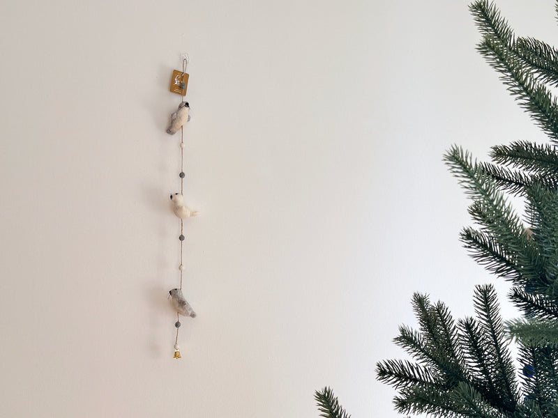 The felt seal garland hangs against a stark white wall in a calm, minimalist setting, with a Christmas tree gently visible nearby. This charming felt garland features three seals, alternating in light gray and white colors.