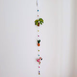 A colorful felt Tropical Trio Flamingo garland hangs vertically against a plain white wall. It features vibrant felt shapes including a green palm tree with a brown trunk, an orange pineapple with green leaves, and a pink flamingo with a white heart and tan legs. These shapes are interspersed with light blue, pink, yellow, and green felt balls, all connected by a fine string, with a copper bell dangling at the bottom.