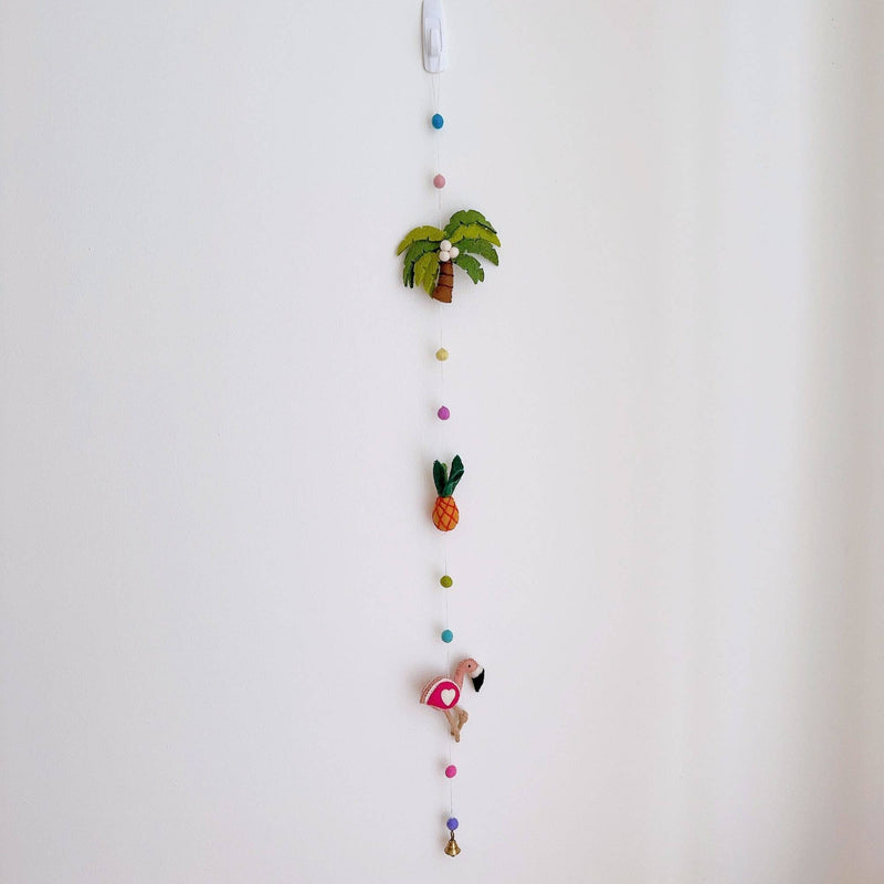A colorful felt Tropical Trio Flamingo garland hangs vertically against a plain white wall. It features vibrant felt shapes including a green palm tree with a brown trunk, an orange pineapple with green leaves, and a pink flamingo with a white heart and tan legs. These shapes are interspersed with light blue, pink, yellow, and green felt balls, all connected by a fine string, with a copper bell dangling at the bottom.