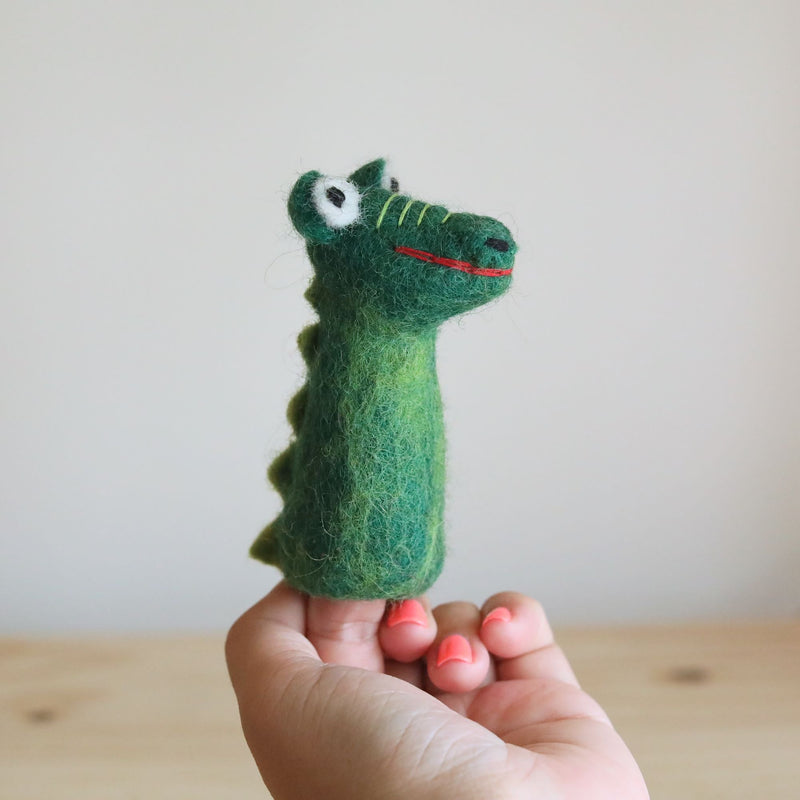 A female hand holds a green handmade wool felt gator finger puppet, showcased against a blurred wooden table background. This charming felt gator features small white eyes, yellow stitching on its snout, and a red smile. Its body has textured scales and small wool felt protrusions running down its back to the tail.