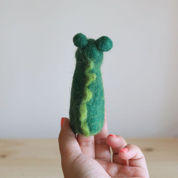 A handcrafted wool felt gator finger puppet is presented upright by a hand with coral-colored nails against a softly lit, muted background. The charming gator features a dark green body with a lighter green zigzag stripe along its back and two small, round ears. The unique finger puppet, emphasizing its whimsical and tactile nature, rests above a light wooden surface in a calm studio setting.