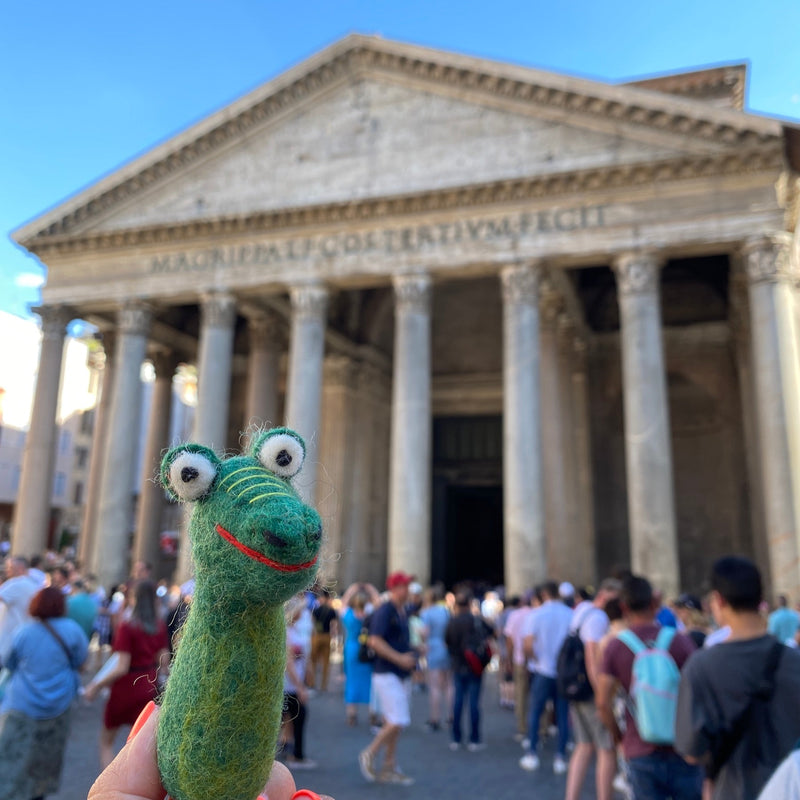 A playful green wool felt gator finger puppet, with a red stitched smile, yellow stripes on its head, and googly eyes, is prominently displayed in a bustling outdoor setting. The handcrafted felt kids toy stands out against the iconic Pantheon under a clear, vibrant sky, exuding a touristy atmosphere.