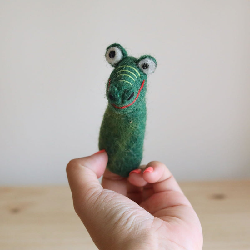 A handmade felt gator finger puppet is held in a female hand over a wooden surface. This green wool felt gator finger puppet features distinct stripes, a detailed mouth, and white and black eyes.