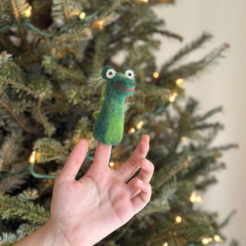 A handcrafted, high-quality wool felt gator finger puppet is playfully presented on a female hand's finger, set against a festive Christmas tree adorned with warm lights. The charming green gator finger puppet features white eyes, a red smile, and textured scales along its back, adding a whimsical touch to the holiday scene.