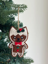 A needle felted gingerbread cat ornament hangs from a green Christmas tree, creating a joyful holiday atmosphere. The light brown cat-shaped ornament features white felt outlining, mimicking gingerbread cookie icing, with a white 'frosting' on its head. It has a red and green holly accent, stitched black eyes, mouth, white whiskers, a red bow tie, and small red and green felt dots on its body.