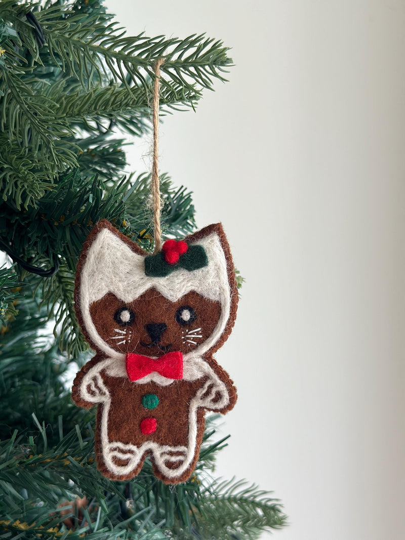 A needle felted gingerbread cat ornament hangs from a green Christmas tree, creating a joyful holiday atmosphere. The light brown cat-shaped ornament features white felt outlining, mimicking gingerbread cookie icing, with a white 'frosting' on its head. It has a red and green holly accent, stitched black eyes, mouth, white whiskers, a red bow tie, and small red and green felt dots on its body.
