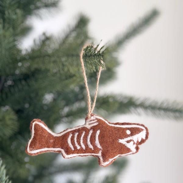 A charming Gingerbread Baby Shark wool felt ornament hangs from a pine branch on a Christmas tree. This unique Christmas ornament is crafted from rich brown wool felt, detailed with white stitching that mimics icing on a gingerbread cookie. A jute string is attached for hanging, evoking a sweet and whimsical holiday mood.