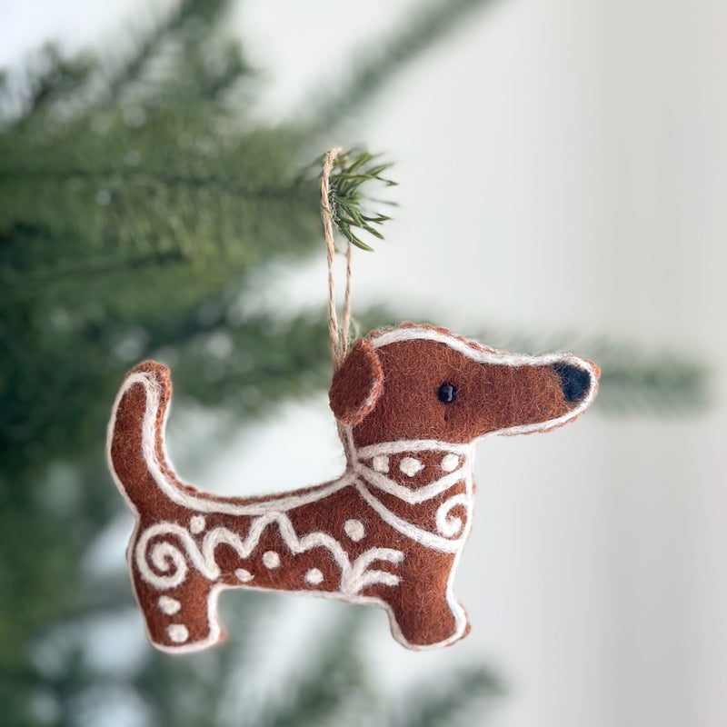 A felt gingerbread dachshund dog ornament hangs festively on a Christmas tree, set against blurred green foliage and a soft white wall. This unique Christmas ornament is predominantly brown, decorated with white swirls and dots resembling gingerbread icing. The charming dachshund has a small black nose and simple black eyes. A jute string allows easy hanging on the tree.
