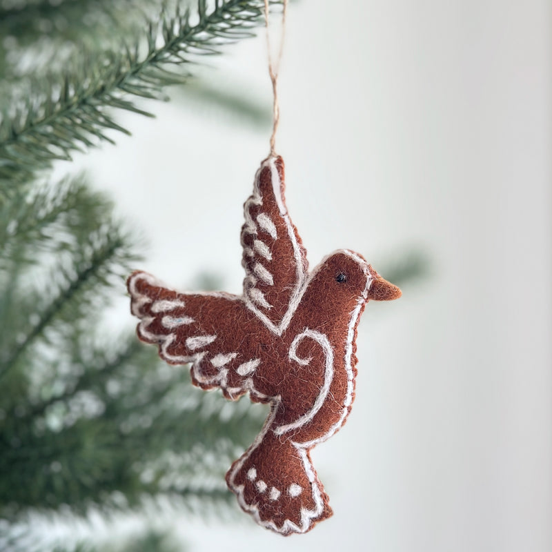 A handcrafted felt gingerbread dove ornament hangs gracefully from a green Christmas tree branch, evoking a cozy holiday atmosphere. The charming brown felt dove features white felt accents that resemble frosting on a gingerbread cookie, highlighting its whimsical design.