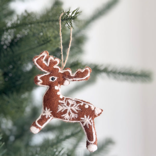 A wool felt reindeer ornament hangs gracefully from a vibrant green Christmas tree branch. This handmade Gingerbread reindeer ornament is crafted from toasty brown wool felt and features playful white stitching, embodying a classic cookie-inspired theme. A jute string suspends it against a soft, blurred white background, evoking holiday cheer.