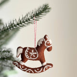 A needle felted gingerbread rocking horse ornament hangs from a Christmas tree branch, evoking a festive mood. This unique Christmas ornament is a rich brown with white details resembling icing. It features decorative swirls on its base and a star on the saddle, suspended by a red and white cotton string.