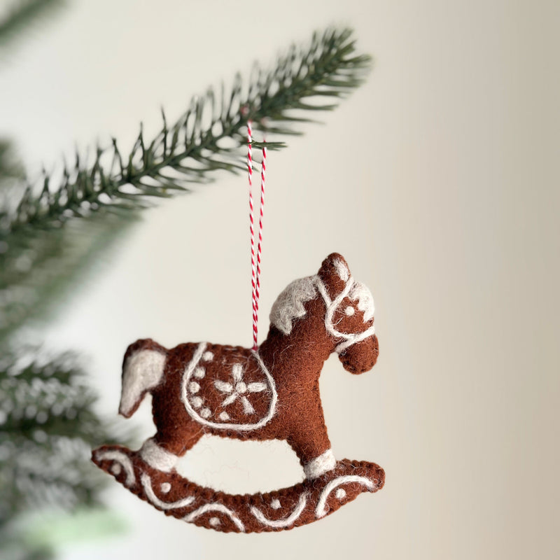 A needle felted gingerbread rocking horse ornament hangs from a Christmas tree branch, evoking a festive mood. This unique Christmas ornament is a rich brown with white details resembling icing. It features decorative swirls on its base and a star on the saddle, suspended by a red and white cotton string.