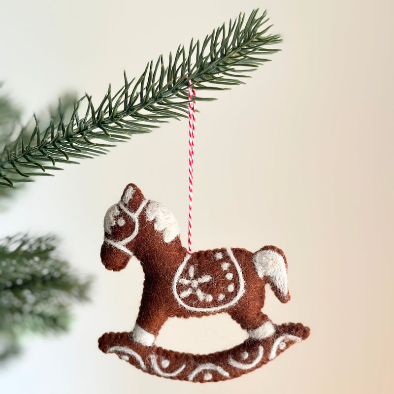 A charming wool felt gingerbread rocking horse ornament hangs from a green Christmas tree branch. This brown rocking horse features white needle-felted details, including a saddle design, mane, and base trim. A red and white cotton string is attached for easy hanging.