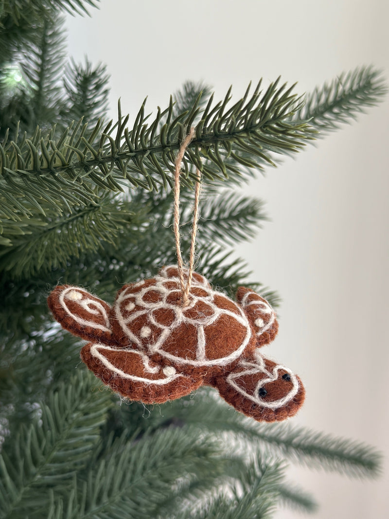 A warm brown wool felt turtle ornament, part of the Felt Gingerbread Collection, hangs on a festive green Christmas tree. This unique Christmas ornament features white decorative stitching in geometric patterns on its shell, head, and flippers, resembling a gingerbread cookie. Small black bead eyes and a jute string for hanging complete the design.