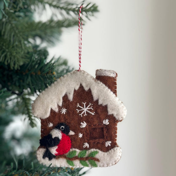 A handmade wool felt gingerbread house with woodpecker Christmas ornament hangs nestled among lush green Christmas tree branches. This charming 3D decoration features a brown felt house with white snow-like trim, a chimney, and stitched window details. A vibrant red, black, and white needle felted woodpecker perches at its base, adding a whimsical touch.