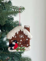 A Felt Gingerbread House with Woodpecker Ornament hangs from a green Christmas tree branch against a white background. This handmade wool felt ornament features a charming gingerbread house with a chimney, snow-like roof, and embroidered snowflakes. A woodpecker is perched on a branch near the bottom of the house. It is suspended by a red and white cotton string.