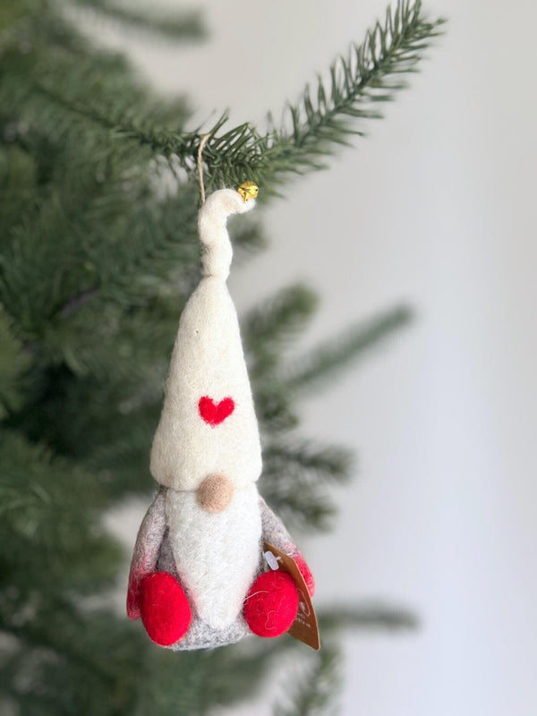 A delightful needle felted gnome Christmas ornament hangs on a green Christmas tree branch. This gnome ornament features a white hat adorned with a red heart, a curly tip with a golden jingle bell, a light brown nose, a white beard, a grey body, and red pompom feet. A small brown tag is attached to one of its feet.