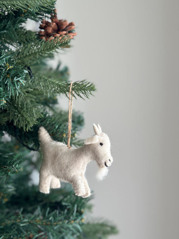 A soft, off-white felt goat ornament, suspended by a jute string, hangs on a vibrant green Christmas tree. The felt goat ornament features a fluffy beard and perky ears, lending a rustic and warm feel to the festive tree decorated with needles and a pine cone.