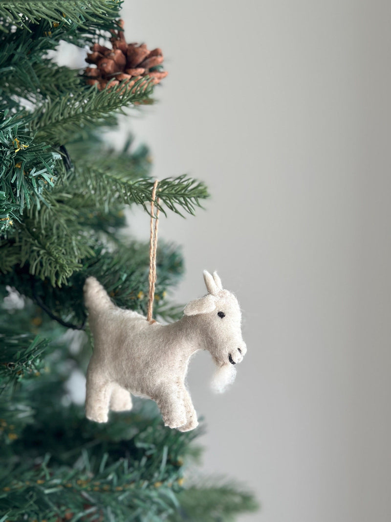 A soft, off-white felt goat ornament, suspended by a jute string, hangs on a vibrant green Christmas tree. The felt goat ornament features a fluffy beard and perky ears, lending a rustic and warm feel to the festive tree decorated with needles and a pine cone.