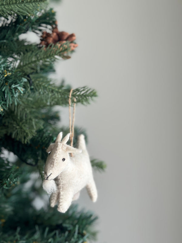 A needle felted goat ornament hangs gracefully from a vibrant green Christmas tree. The creamy, off-white wool felt goat features small dark eyes and a fluffy beard. Its needle-felted texture and attached jute string highlight its handcrafted appeal and festive charm.