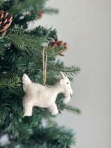 A needle felted goat ornament, featuring a jute string, hangs close-up on a green Christmas tree adorned with pine cones. This off-white goat has a fluffy texture, with small ears, a tiny tail, a beard, and dark eyes, adding a charming, rustic appeal and seasonal warmth to the festive backdrop.
