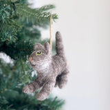 A wool felt gray cat ornament hangs on a green Christmas tree. The handcrafted gray cat features textured wool felt, a small pink nose, white whiskers, and pointy ears. Suspended by a jute string, it adds a cozy, playful touch to the festive decor.