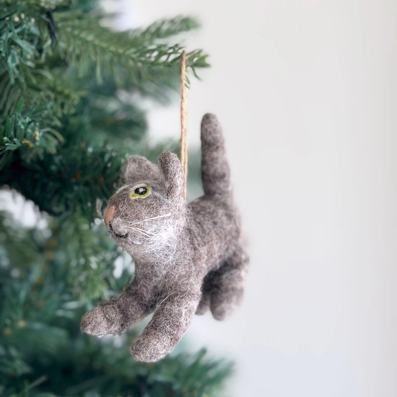 A wool felt gray cat ornament hangs on a green Christmas tree. The handcrafted gray cat features textured wool felt, a small pink nose, white whiskers, and pointy ears. Suspended by a jute string, it adds a cozy, playful touch to the festive decor.