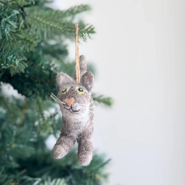 A handmade felt gray cat ornament, featuring a charming gray body with small ears, a peach-colored nose, and delicate white whiskers, hangs from a jute string on a Christmas tree. This cozy wool felt cat ornament adds a unique, handcrafted appeal to the festive decor.