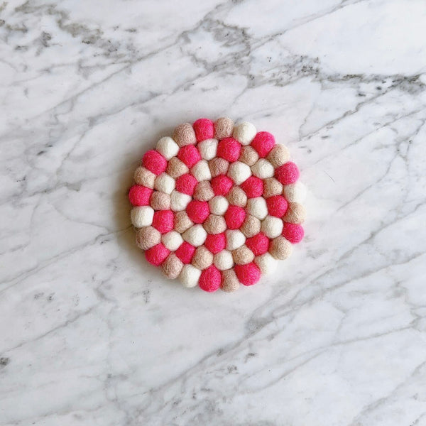 A round 'Happily Ever After' felt coaster, one from a set of four, rests on a white marble surface with subtle grey veining. This handcrafted wool felt coaster, ethically made from 100% organic lamb wool by Nepalese female artisans, features tightly hand-stitched half-inch felt balls. Its vibrant design showcases alternating shades of bright pink, light beige, and white, creating a textured and colorful surface perfect for home decor.