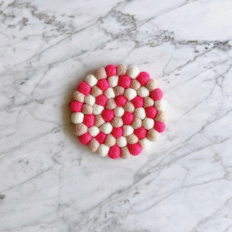 A round 'Happily Ever After' felt coaster, one from a set of four, rests on a white marble surface with subtle grey veining. This handcrafted wool felt coaster, ethically made from 100% organic lamb wool by Nepalese female artisans, features tightly hand-stitched half-inch felt balls. Its vibrant design showcases alternating shades of bright pink, light beige, and white, creating a textured and colorful surface perfect for home decor.
