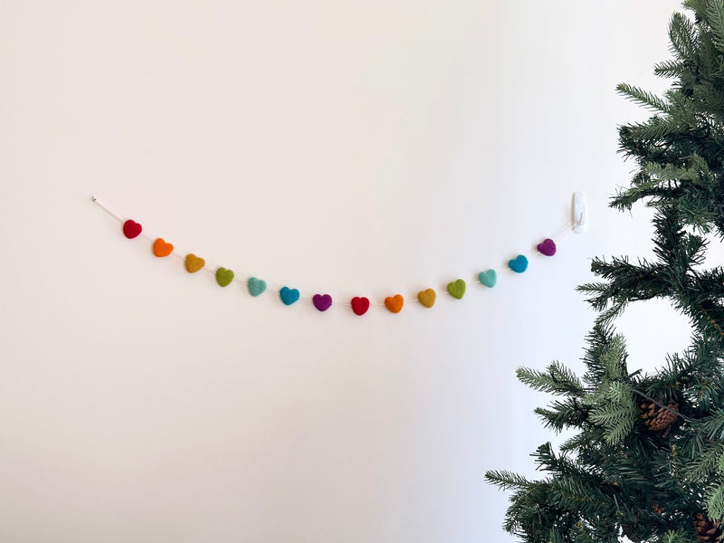 A cheerful felt garland, featuring seven rainbow-hued pastel stars strung on a cotton thread, is displayed against a white wall, secured by a clear hook. An artificial pine tree stands to its side, enhancing the festive atmosphere.