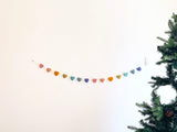 A felt star garland is strung horizontally against a stark white wall, with a partial Christmas tree visible on the right. The garland features an array of uniformly shaped felt stars in pastel colors including pink, peach, mustard, green, turquoise, lilac, and navy. Suspended by a delicate cotton thread, it adds a playful, charming accent, hinting at festive decor.