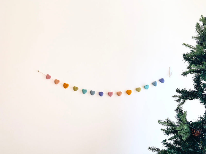 A felt star garland is strung horizontally against a stark white wall, with a partial Christmas tree visible on the right. The garland features an array of uniformly shaped felt stars in pastel colors including pink, peach, mustard, green, turquoise, lilac, and navy. Suspended by a delicate cotton thread, it adds a playful, charming accent, hinting at festive decor.