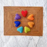 A vibrant set of seven rainbow felt heart ornaments is arranged in a circular pattern on a brown woven mat, set against a marble background. These cheerful felt heart ornaments include red, orange, yellow, light green, light blue, dark blue, and purple hearts. Each soft, textured heart is crafted from felt and suspended with a jute string, creating a handcrafted feel.