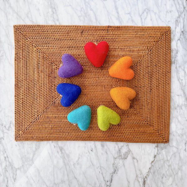 A vibrant set of seven rainbow felt heart ornaments is arranged in a circular pattern on a brown woven mat, set against a marble background. These cheerful felt heart ornaments include red, orange, yellow, light green, light blue, dark blue, and purple hearts. Each soft, textured heart is crafted from felt and suspended with a jute string, creating a handcrafted feel.