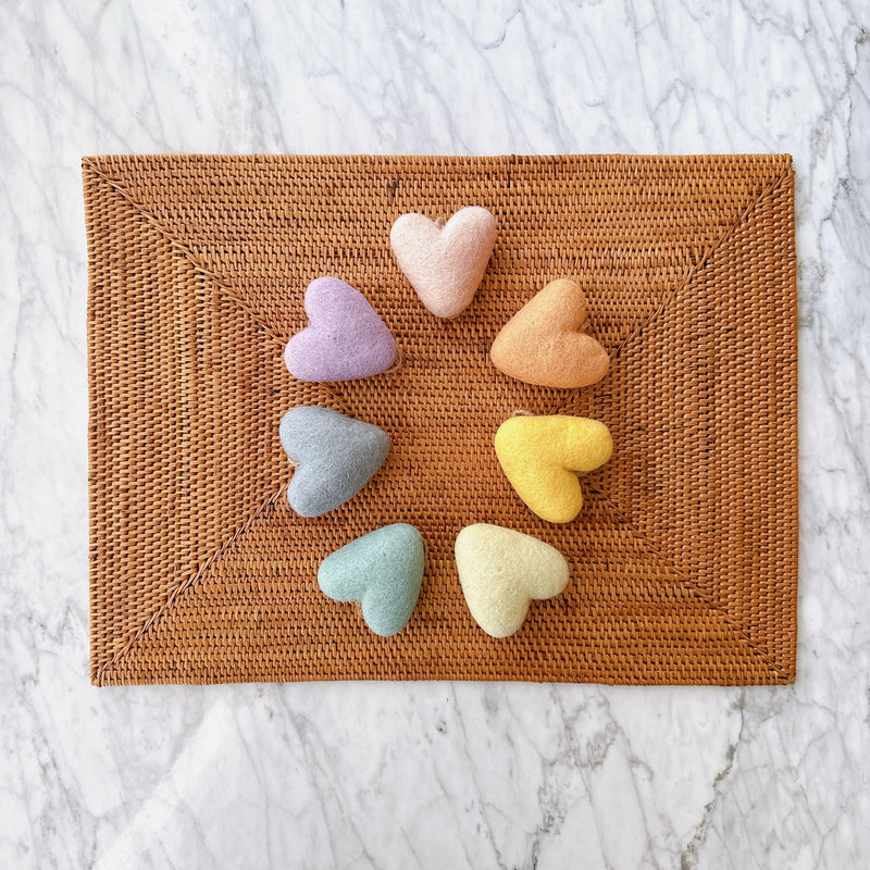 A Rainbow Set of 7 felt heart ornaments is arranged in a circle on a brown woven placemat, resting on a white marble surface. These felt heart ornaments are soft and fluffy, each featuring a unique pastel color, creating a vibrant rainbow palette. Each heart is strung with twine for easy hanging, adding a calm and decorative touch.