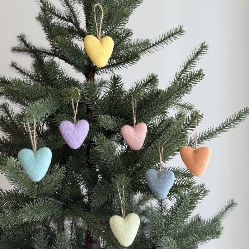 Seven wool felt heart ornaments from a rainbow set are closely displayed on a lush Christmas tree. Each unique handmade felt ornament showcases a soft pastel hue, including yellow, lavender, light pink, light blue, light gray blue, peach, and light green, suspended by rustic jute string. The handcrafted felt ornaments create a festive yet minimalist display, their soft texture contrasting with the tree's smooth needles.