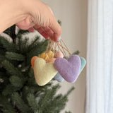 This cheerful set of seven rainbow felt heart ornaments is delicately displayed in a female hand, creating a vibrant contrast against a lush green Christmas tree. Each pastel-colored felt heart, featuring a soft texture in hues of peach, lemon yellow, lavender, mint green, and grey, is suspended by a rustic jute string.