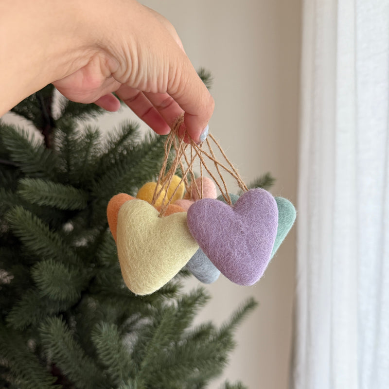 This cheerful set of seven rainbow felt heart ornaments is delicately displayed in a female hand, creating a vibrant contrast against a lush green Christmas tree. Each pastel-colored felt heart, featuring a soft texture in hues of peach, lemon yellow, lavender, mint green, and grey, is suspended by a rustic jute string.