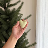 A pastel green felt heart ornament, part of a Rainbow Felt Heart Ornament set, is gently held by a female hand near an evergreen Christmas tree. This soft, textured wool felt ornament is suspended by a jute string, creating a warm, festive atmosphere against a blurred white curtain background.