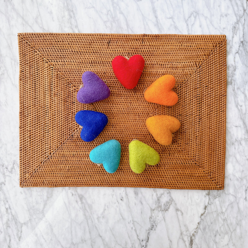 Rainbow felt heart ornaments, a set of seven, are arranged in a cheerful circular pattern on a rustic woven mat, set against a bright marble background. Each handcrafted felt heart is a different vibrant color of the rainbow, featuring a slightly textured surface and a natural twine loop for easy hanging.