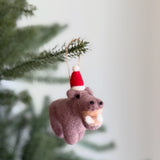 A felt hippo ornament hangs from a blurred green Christmas tree branch against a soft, neutral backdrop. This chunky, light purple needle-felted wool hippo wears a small red Santa hat with a white pompom. It features small black eyes and large white felt teeth, with a jute string attached for hanging.